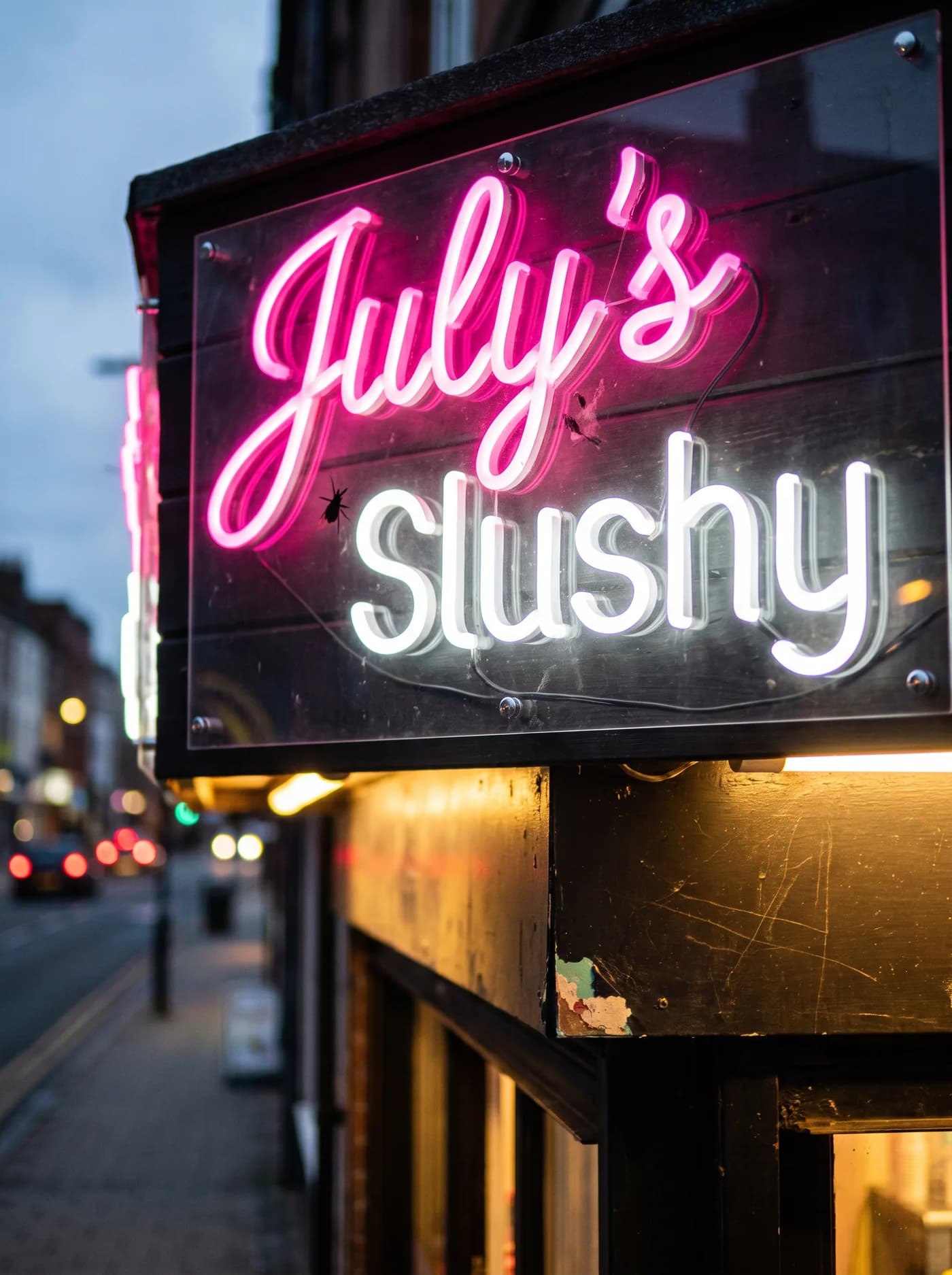 Close-up of a two-line neon sign: pink lettering above white lettering, mounted on an acrylic panel above a shop entrance