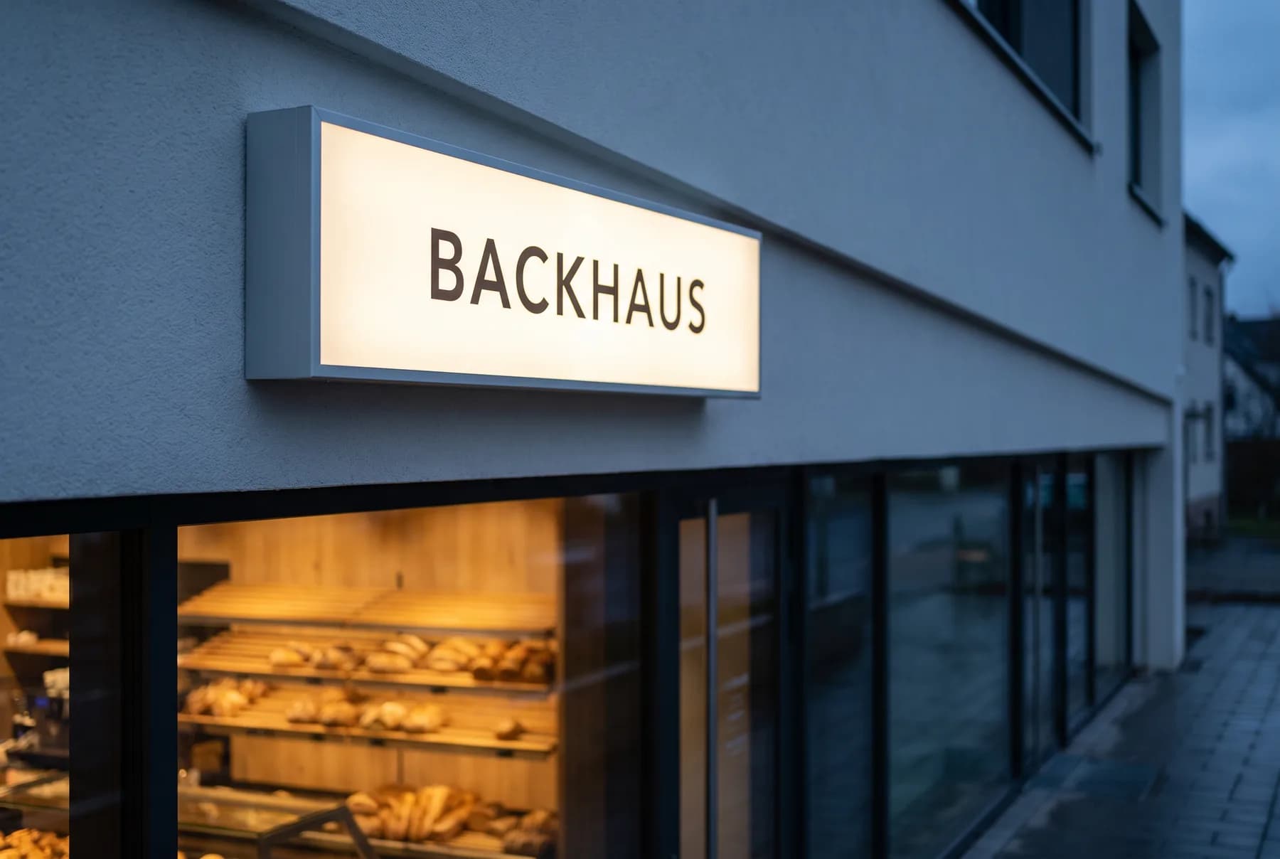 Rechteckiger Leuchtkasten mit bedruckter, gleichmäßig hinterleuchteter Front über dem Eingang einer Bäckerei in der Dämmerung